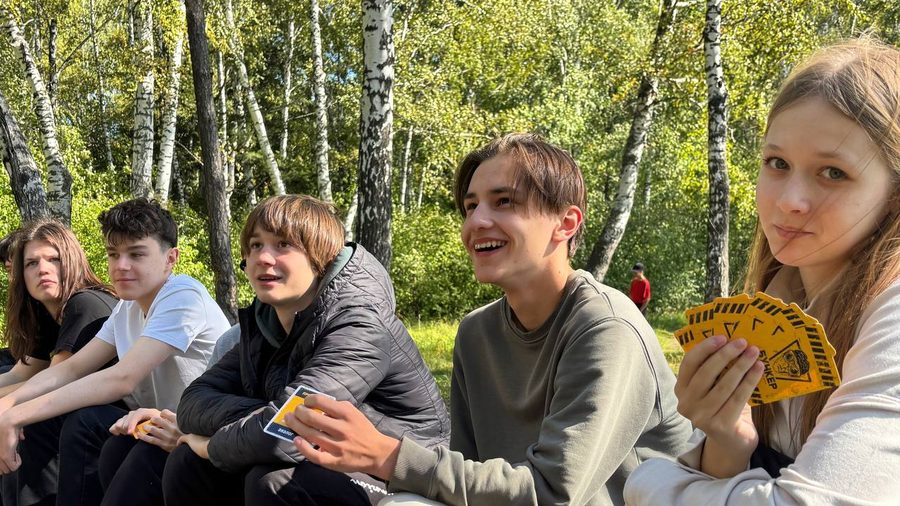 Школьники сидят на поляне в берёзовом лесу и играют в настольную игру во время поездки на природу